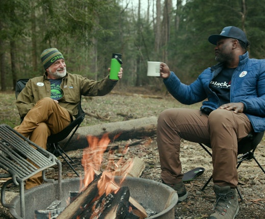 Two men cheers their drinks around a campfire while wearing Oboz.