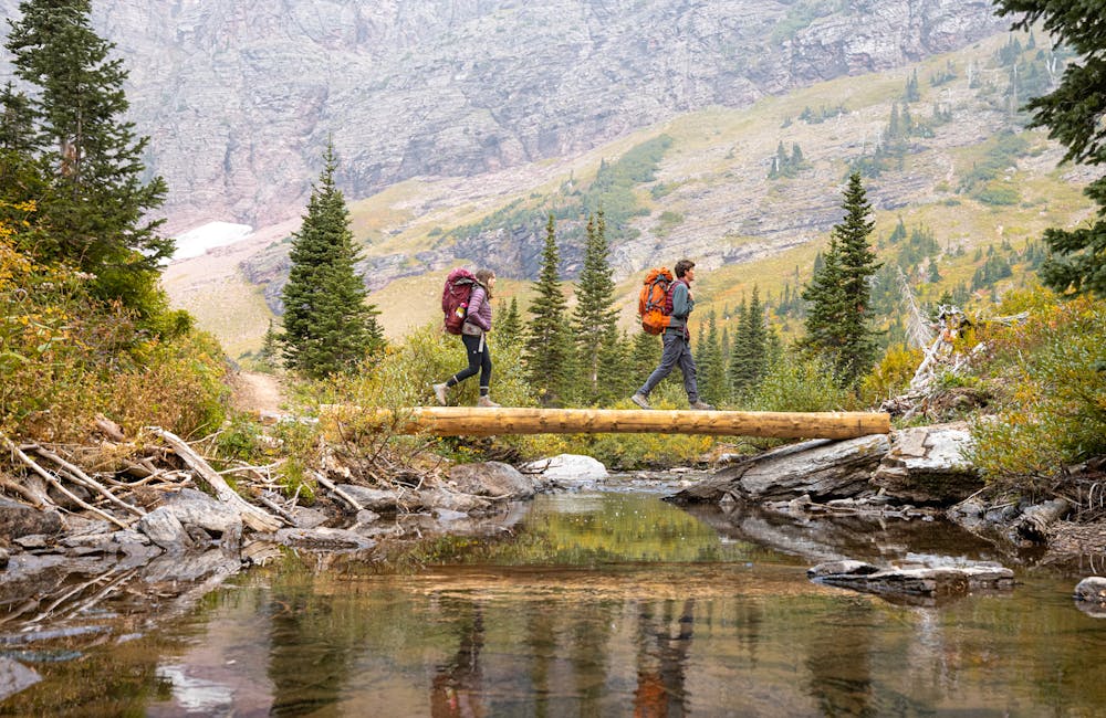 Two hikers cross over a mountain creek in Oboz hiking boots while backpacking in Montana.