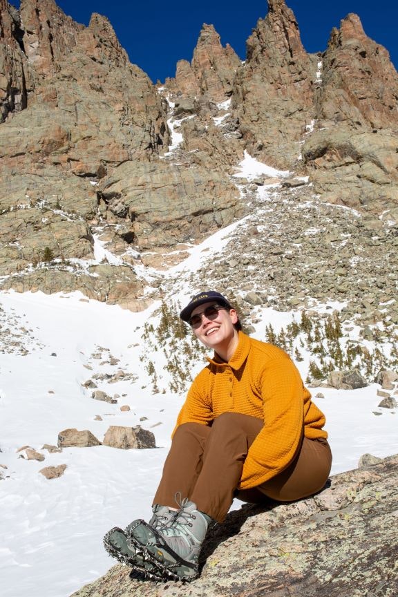 Kylie Yang sits on a snowy boulder in the mountains wearing her Oboz Bangtail winter hiking shoes. 