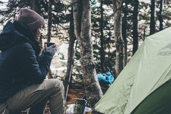 Woman camping in the woods and drinking coffee in the morning by her tent.