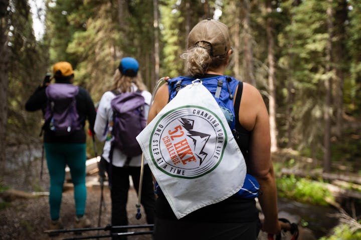 Participants of the 52 Hike Challenge, hike on a forest trail in Montana's Hyalite Valley. The 52 Hike Challenge flag is featured on a woman's pack.