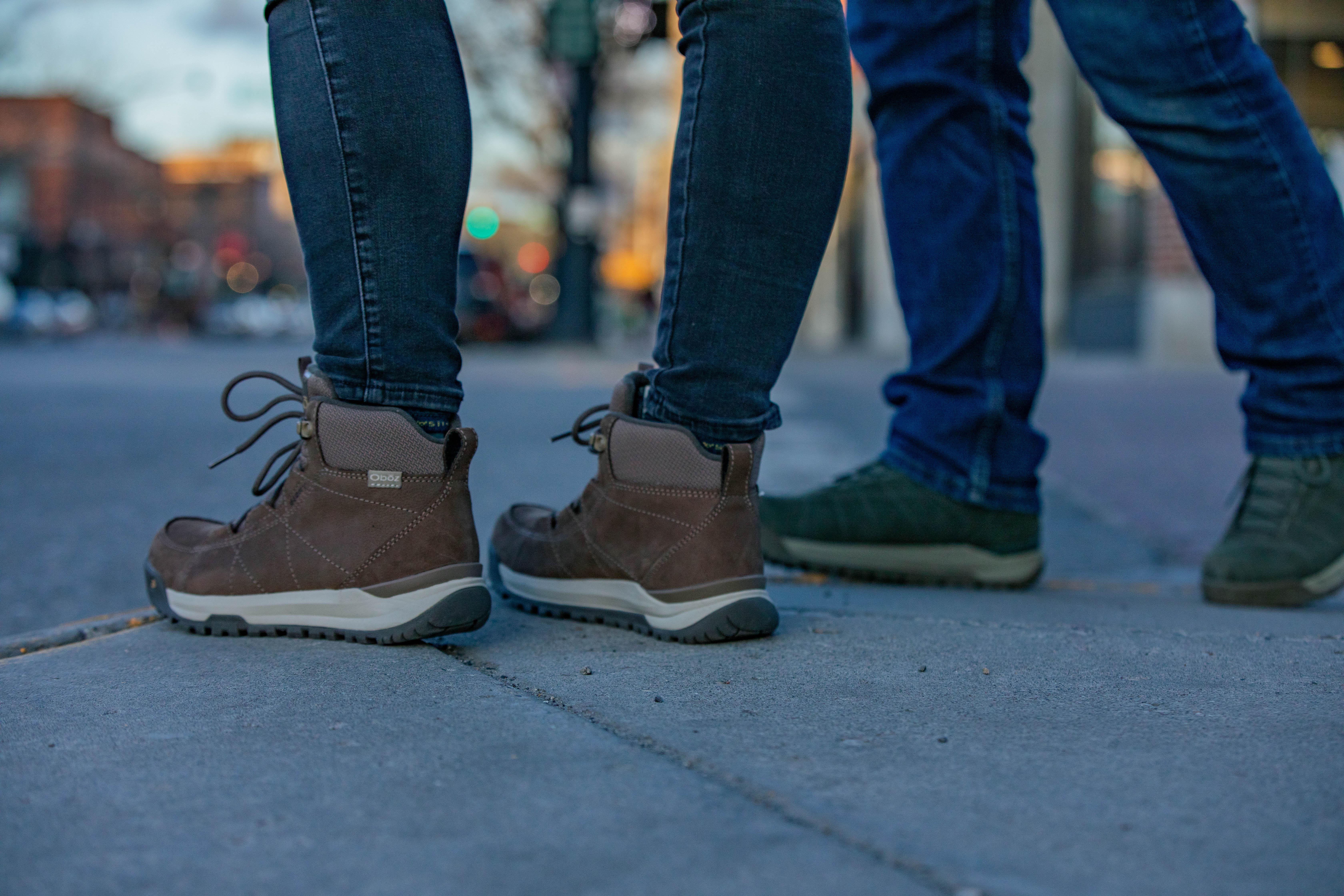 Two people in Oboz winter boots wait at a crosswalk cross the street.