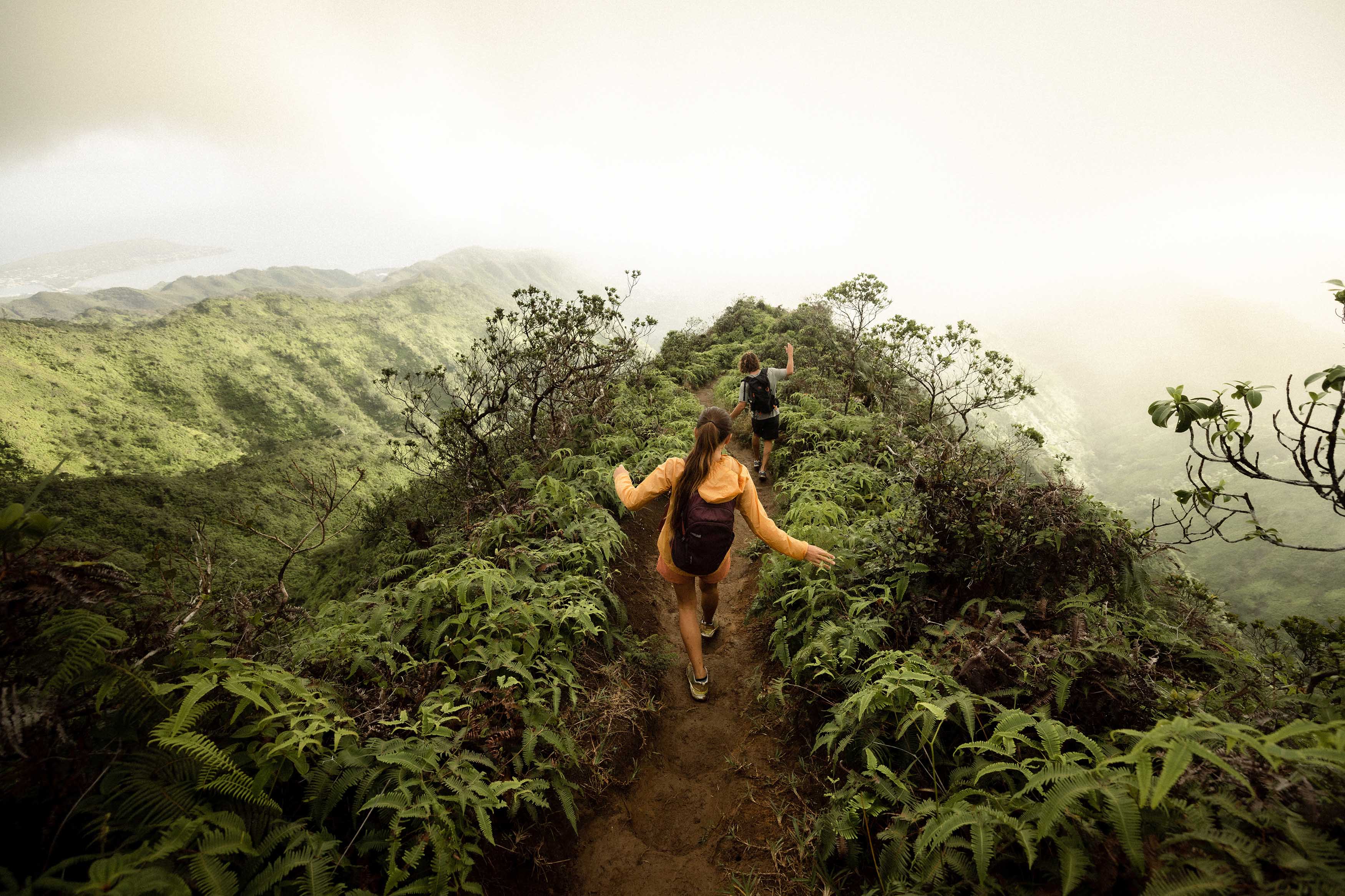 Two hikers navigating a muddy trail on a ridge of a tropical rainforest while wearing Oboz hiking shoes.