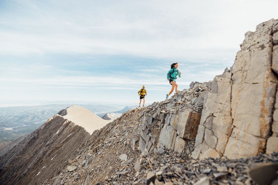 Two people run up a rocky ridge in a blue and yellow jacket wearing Oboz Katabatic trail shoes. 