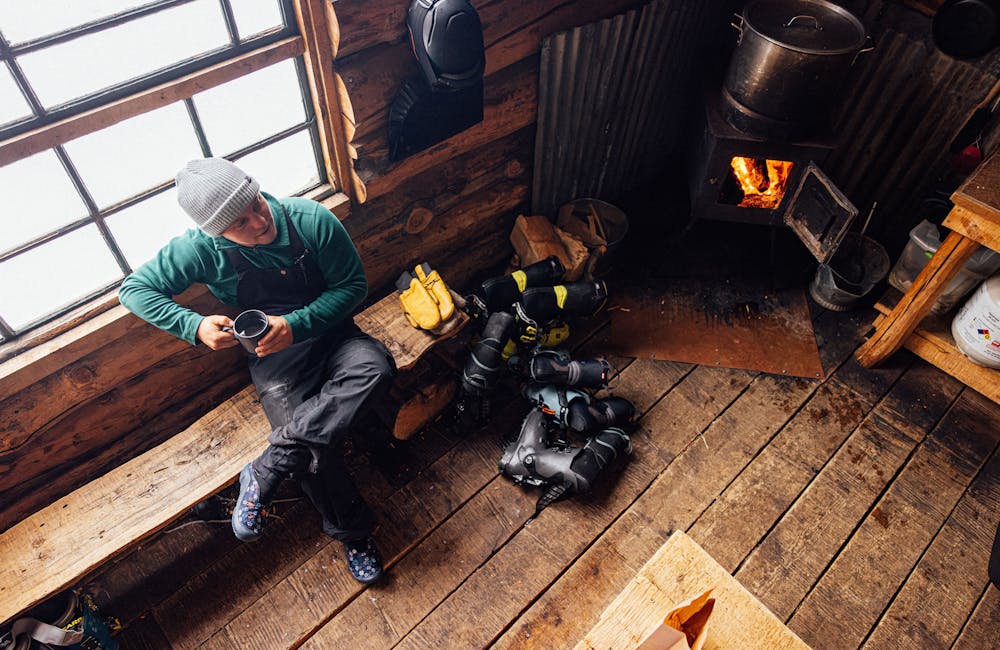 A man drinks coffee in front of a window wearing the Oboz and Skida Whakatā Puffy