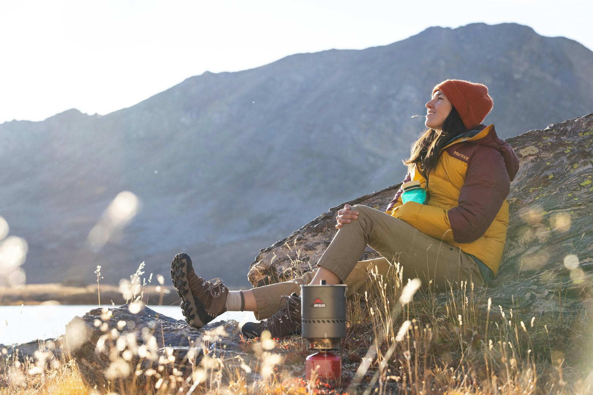 Person in puffer jacket, hiking pants, and Oboz hiking shoes sits on a rock in the wilderness.