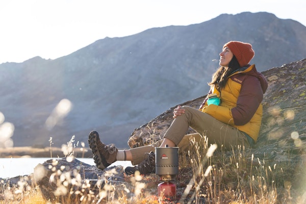 Person in puffer jacket, hiking pants, and Oboz hiking shoes sits on a rock in the wilderness.
