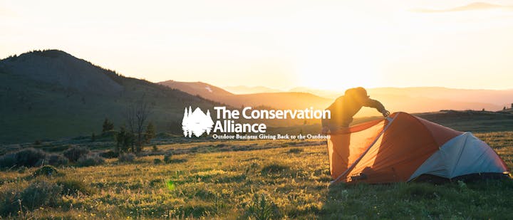 Person in a grassy field zipping up a tent during sunset
