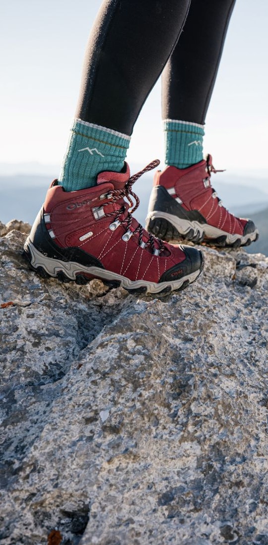 Person wearing Oboz Bridger hiking boot in Rio Red with blue socks on a rocky mountain top.