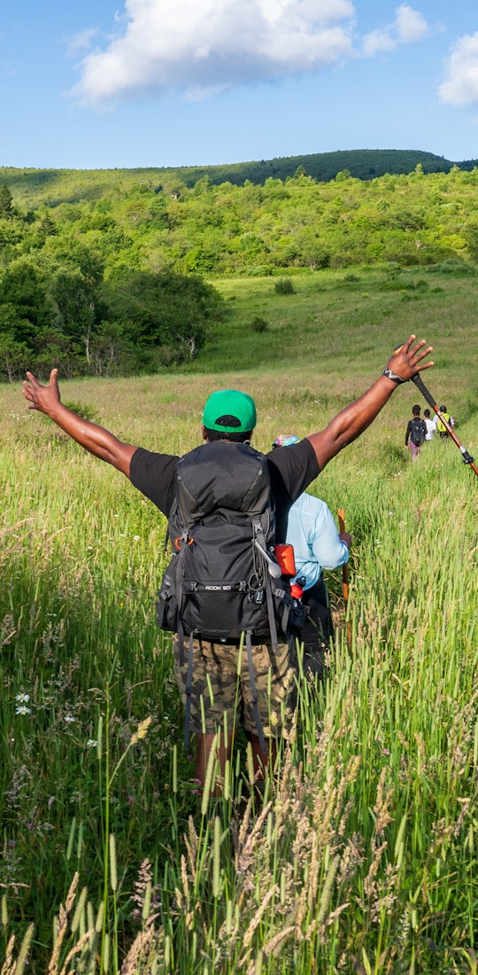Hiker is raising hands victoriously while walking through a grassy plain.