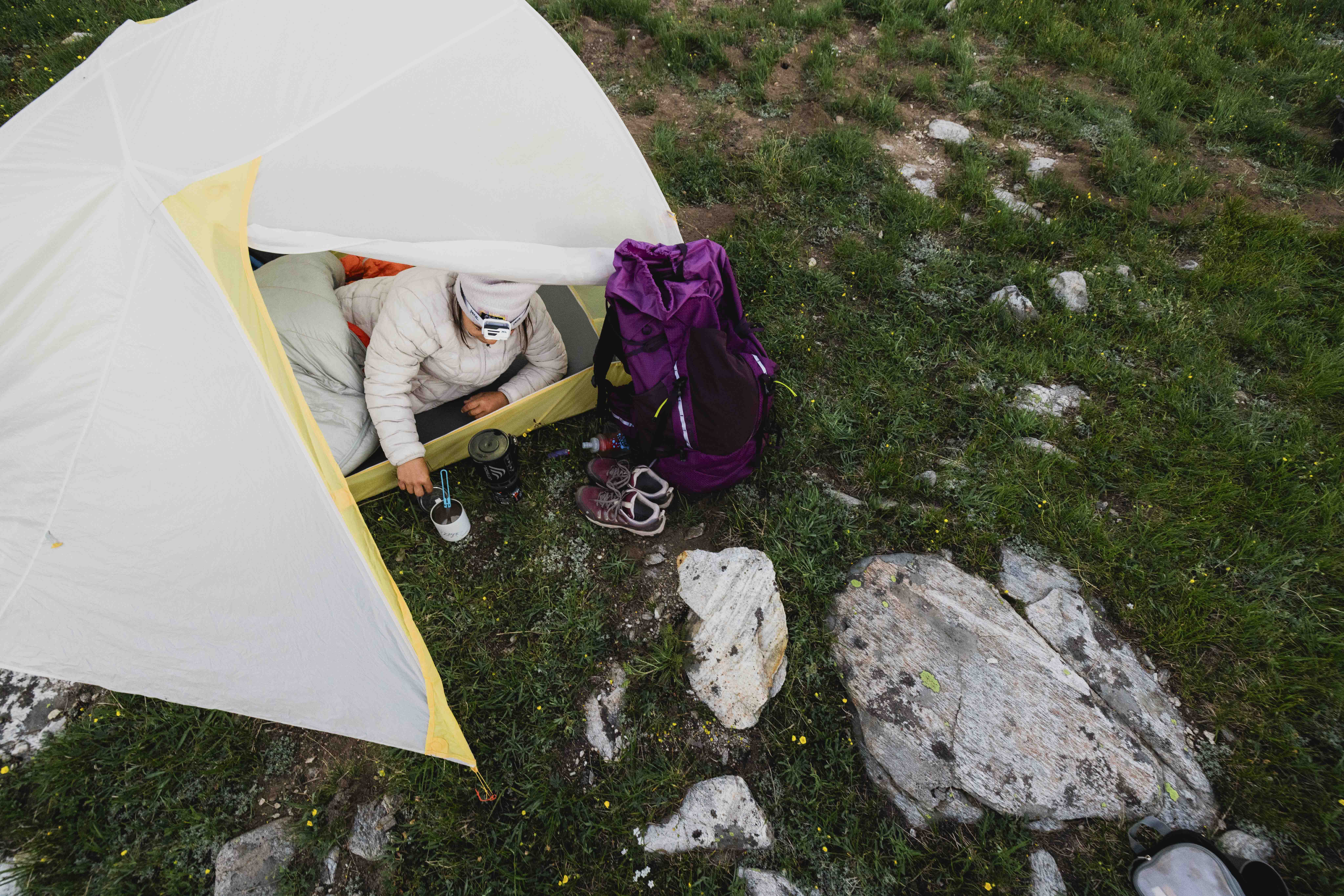 A person drinking coffee in a tent with Oboz hiking boots and a backpack outside.