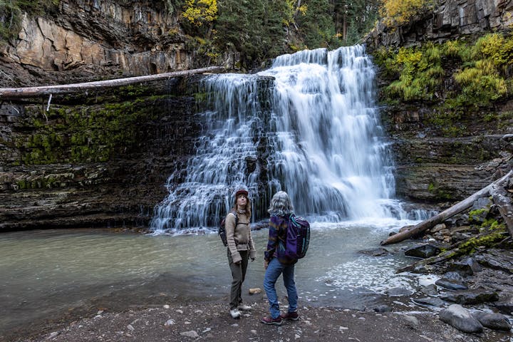 Women stand at the base of a waterfall while on a hike in Montana.