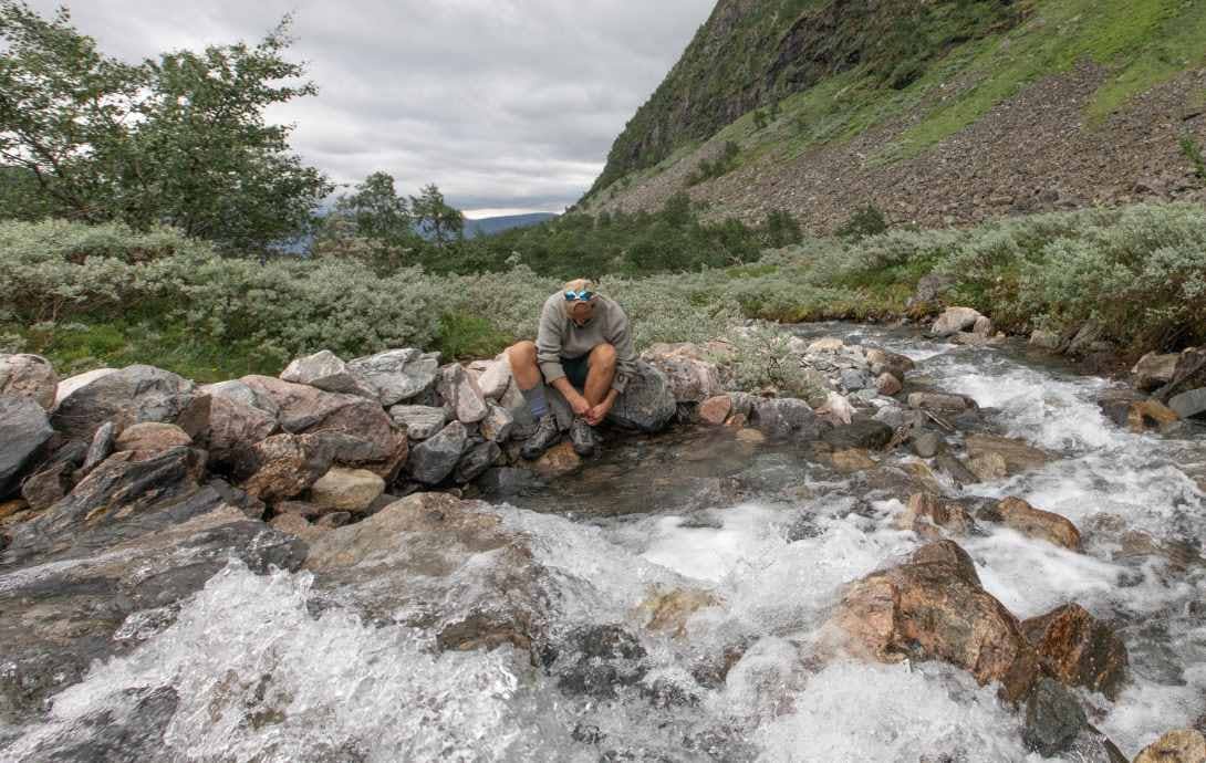 A person ties their shoelaces on their Oboz shoes by a river during a hiking trip.