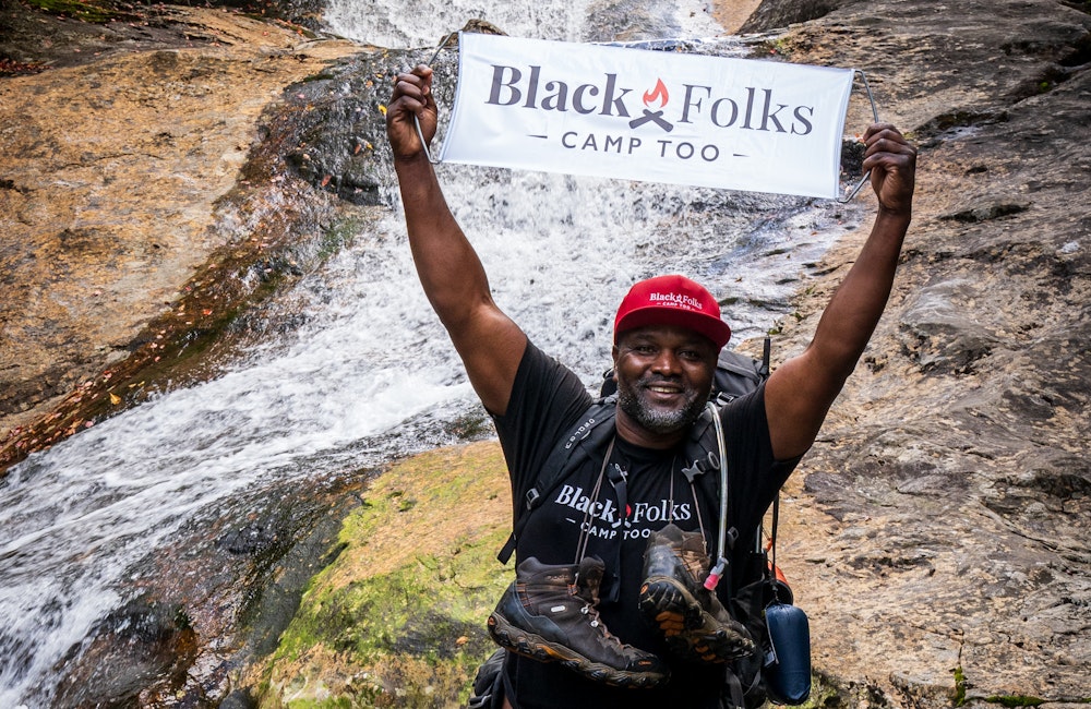 Earl Hunter in front of a waterfall holding up a Black Folks Camp Too sign