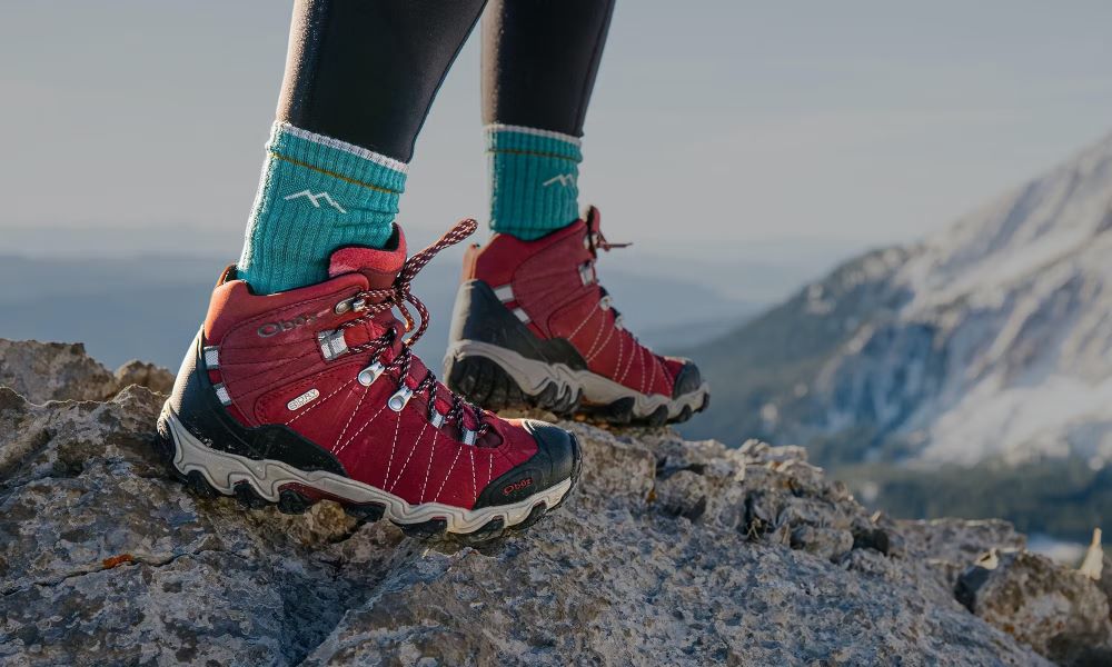 Person wearing Oboz Bridger hiking boot in Rio Red with blue socks on a rocky mountain top.