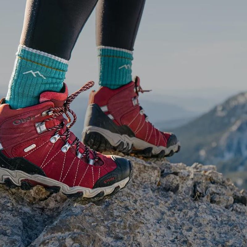 Person wearing Oboz Bridger hiking boot in Rio Red with blue socks on a rocky mountain top.