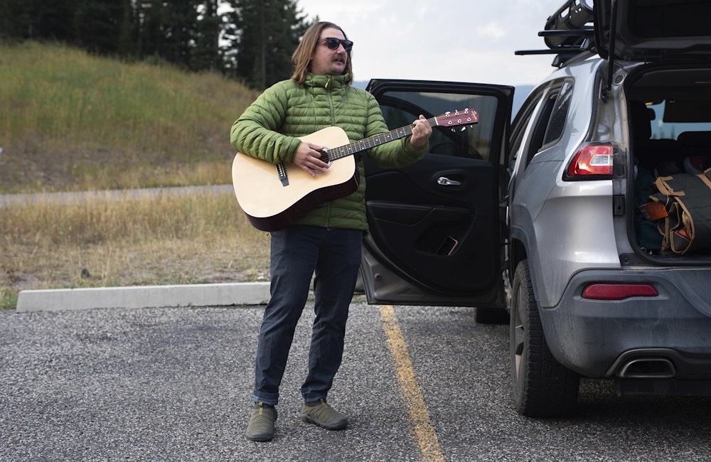 Long haired man playing guitar at Hyalite Lake in Oboz Whakata Puffy slippers.